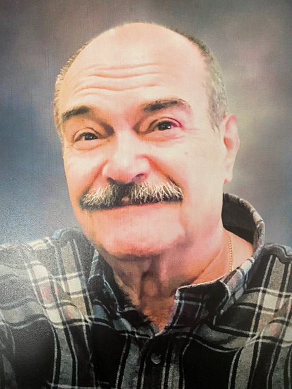 older man with greying mustache and hair smiling with his mouth closed. He is wearing a black and grey plaid shirt. 