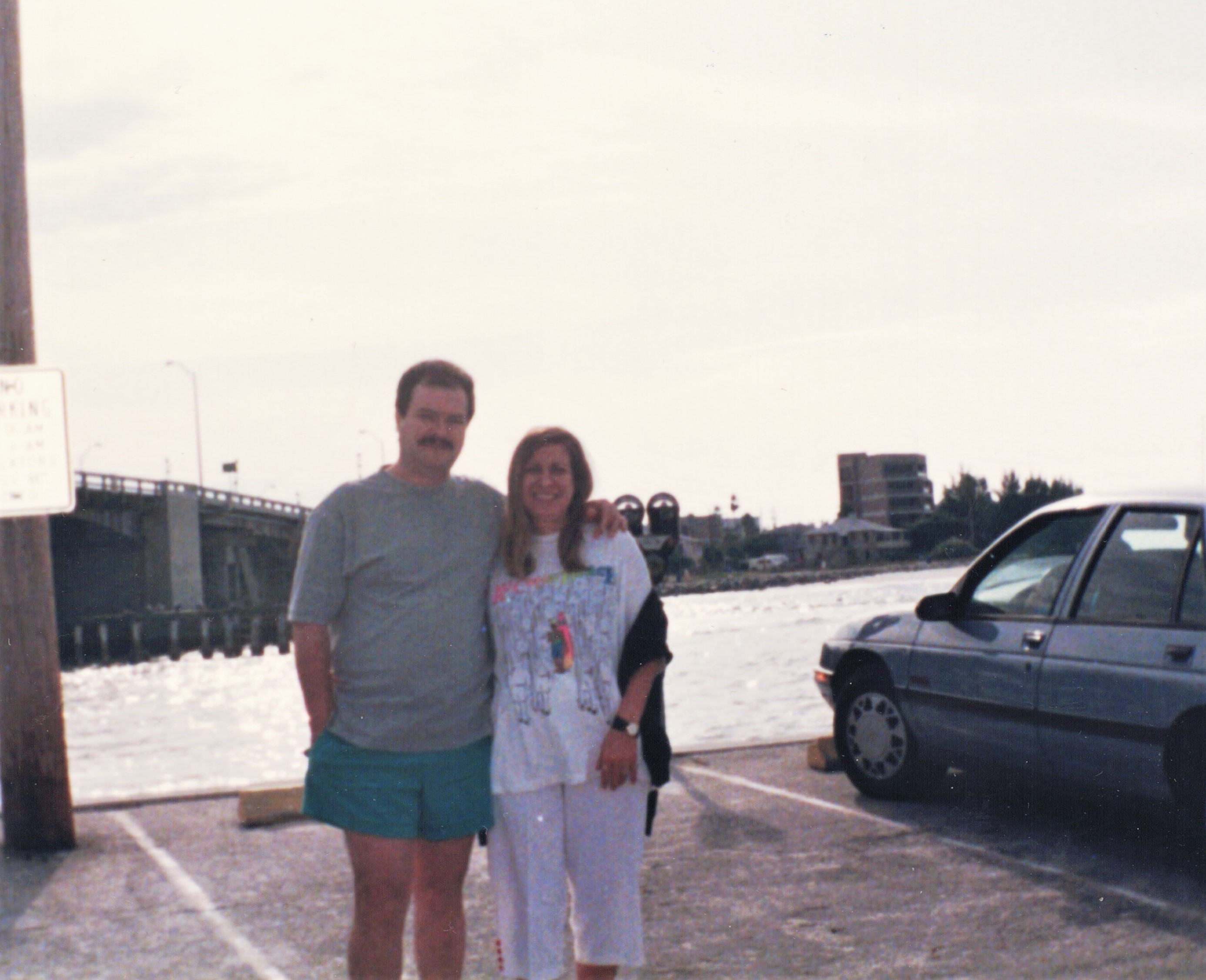 man and woman standing in a parking lot. He is wearing green shorts and a grey tshirt with his arm around the woman. She has on white pants and a white tshirt. 
