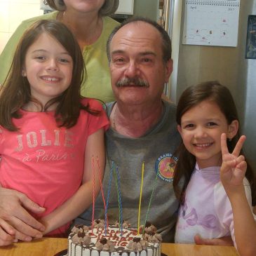 older man sitting with two girls beside him and a women behind him smiling in front of a birthday cake with candles
