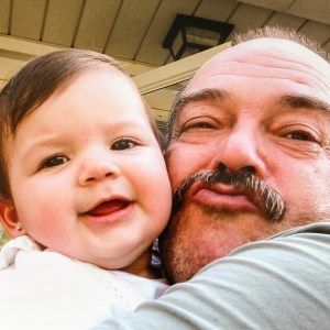 Older man with mustache is cheek-to-cheek with a baby with brown hair and brown eyes. He is puckering his lips for a kiss. 