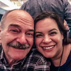 older man with mustache is posing for a selfie with a woman with dark hair. He is wearing a black and burgandy plaid shirt. 